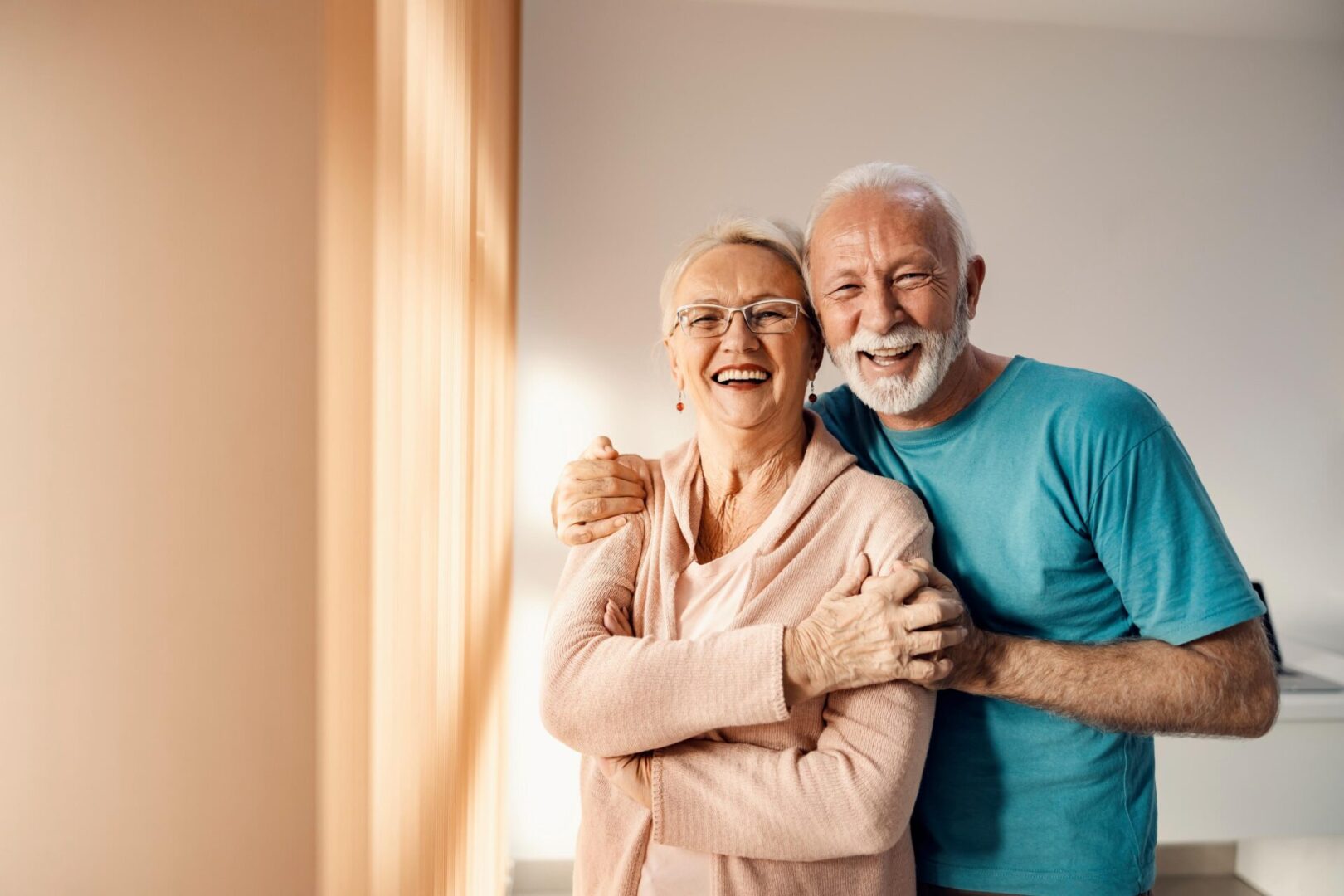 An elderly couple smiling and embracing in a brightly lit room.
