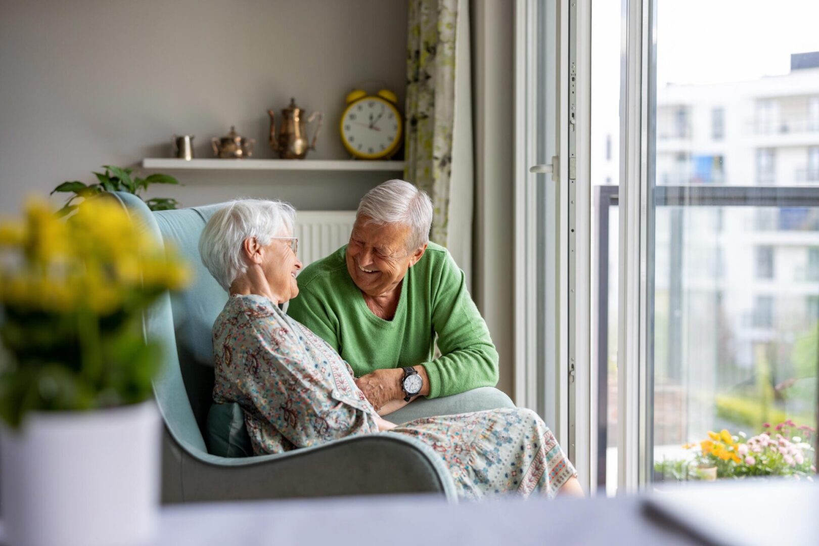 An elderly man and woman sit together in an armchair by a window, smiling at each other. There is a yellow flower in the foreground and a clock on the shelf in the background.
