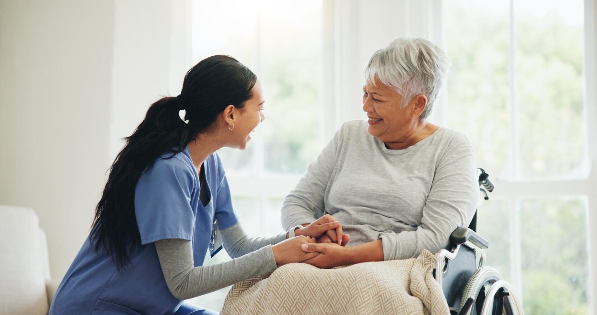 A caregiver in blue scrubs holds hands with an older woman in a wheelchair, both smiling at each other in a bright room.